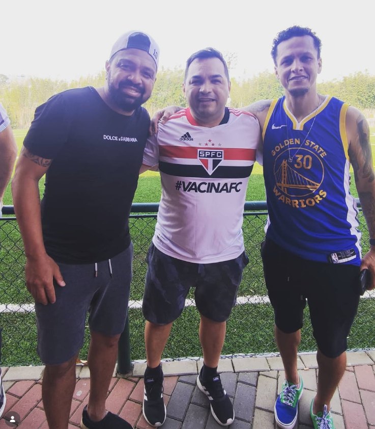 Jura com a camisa do São Paulo FC ao lado de dois amigos, à beira de um campo